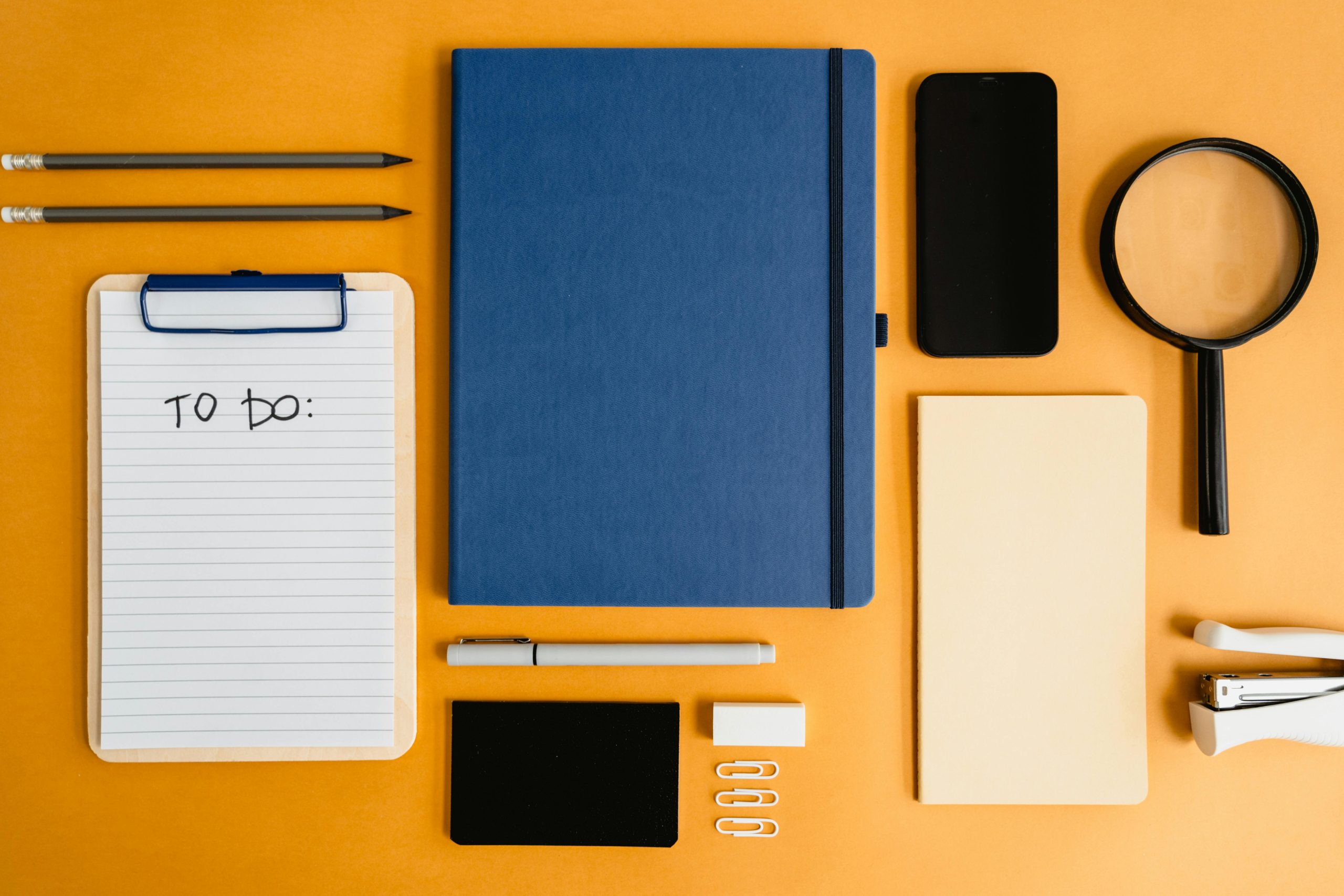 A top view of office stationery including a notebook, to-do list, and magnifying glass arranged on a yellow background.